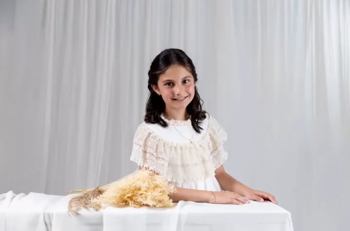 Niña de comunión sentada ante una mesa con cortinas blancas